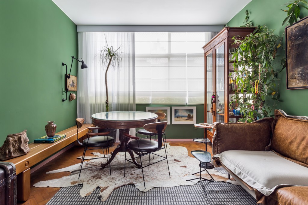 Sala de estar com paredes verdes; mesa de jantar redonda; tapetes; sofá em couro; banco em madeira