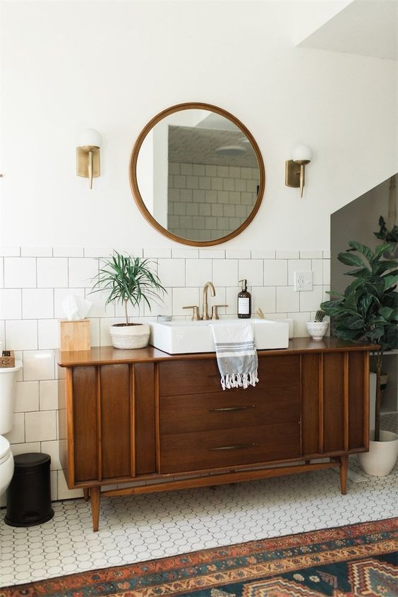 Banheiro com móvel de madeira antigo que faz as vezes de uma bancada para o lavatório. Este que é feito em porcelana branca e com uma torneira moderna, dourada.