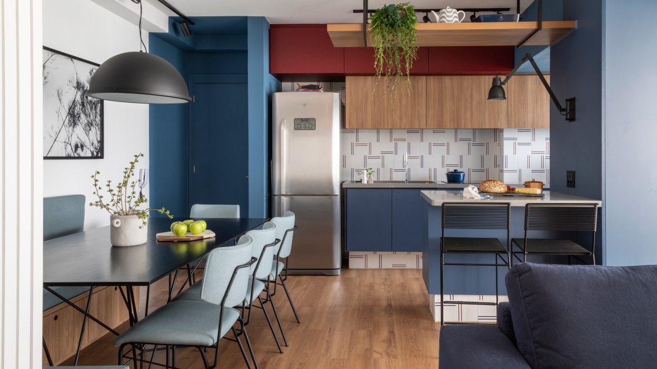 Sala de jantar integrada com cozinha. Móveis desta são azuis e a geladeira de inox. A mesa e cadeiras da sala de jantar são minimalistas. O design como um todo se baseia em linhas e ângulos retos.