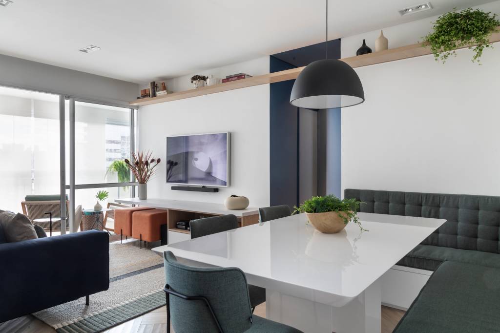 Salas de jantar e estar integradas. Na de jantar, há uma mesa branca sob uma luminária preta em forma de hemisfério. A sala de estar conta com um sofá, dois pufes e uma TV fixada na parede.