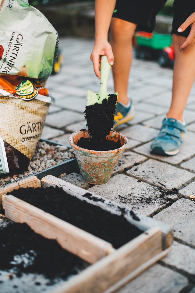 Em uma área externa, uma pessoa deposita terra em um vaso de barro com uma pá para jardinagem