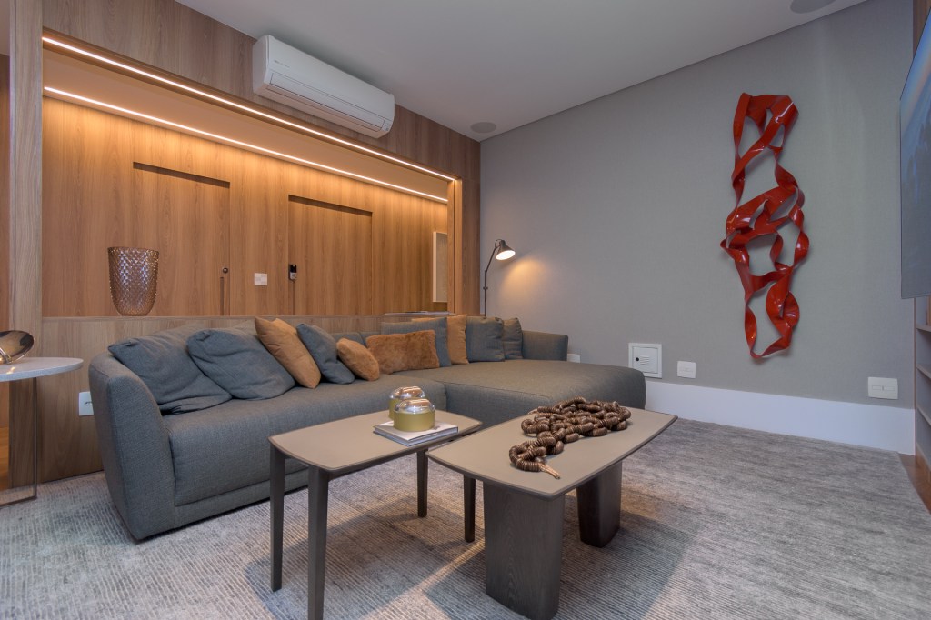 Sala de estar com um grande sofá azul acinzentado. A parede atrás deste móvel é revestida de madeira e guarda fita de led que auxiliam na iluminação do ambiente.
