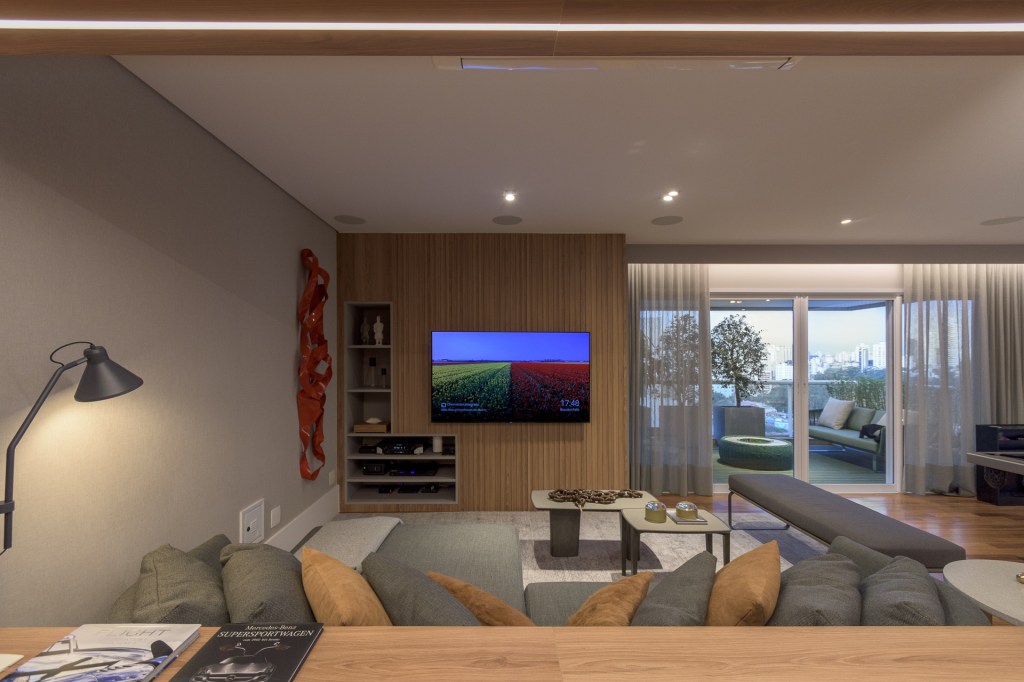 Vista da sala de estar a partir do ponto de vista do sofá. É possível ver uma TV em frente, fixada em uma parede de madeira. No teto, spots de luz iluminam o ambiente.