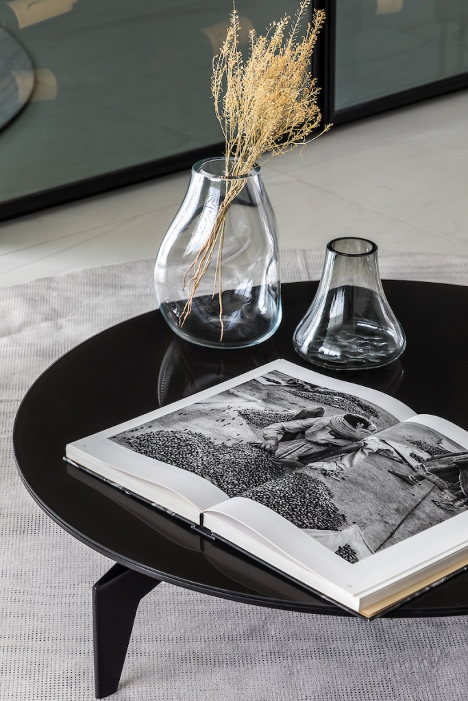 Detalhe de mesa de centro preta, decorada por vasos de vidro em formas sinuosas. Um livro de fotografia aberto compõe a decoração.
