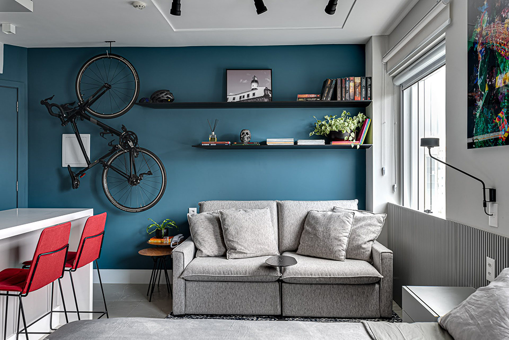 Sala integrada com sofá retrátil, parede azul com prateleiras, bicicleta pendurada