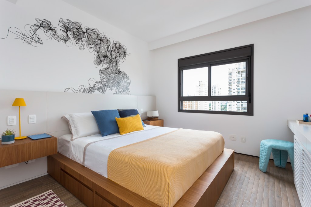 Dormitório com cama e mesinhas laterais de madeira. A parede de cabeceira traz uma ilustração abstrata em linhas pretas. Num canto do quarto está um pufe azul estruturado em tranças com três pés.
