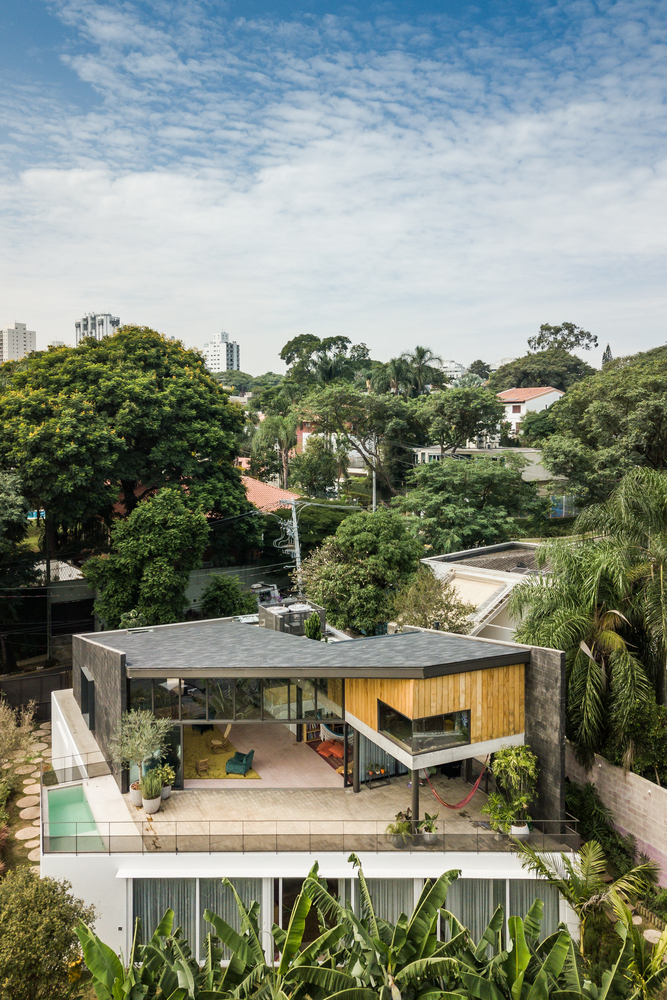 Vista aérea mostra casa com grande varanda e sala de estar no piso superior, em meio à paisagem urbana com bastante vegetação ao redor.