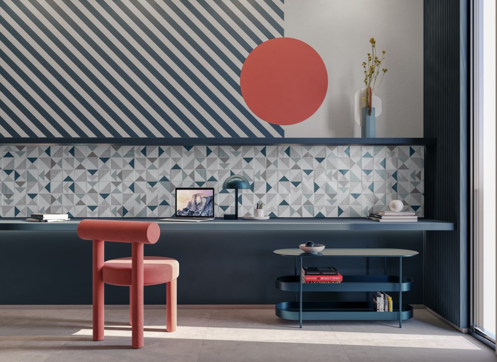 Foto mostra home office com bancada cinza, parede com revestimento cerâmico e pintura artística, cadeira vermelha e mesa de apoio com objetos de escritório.