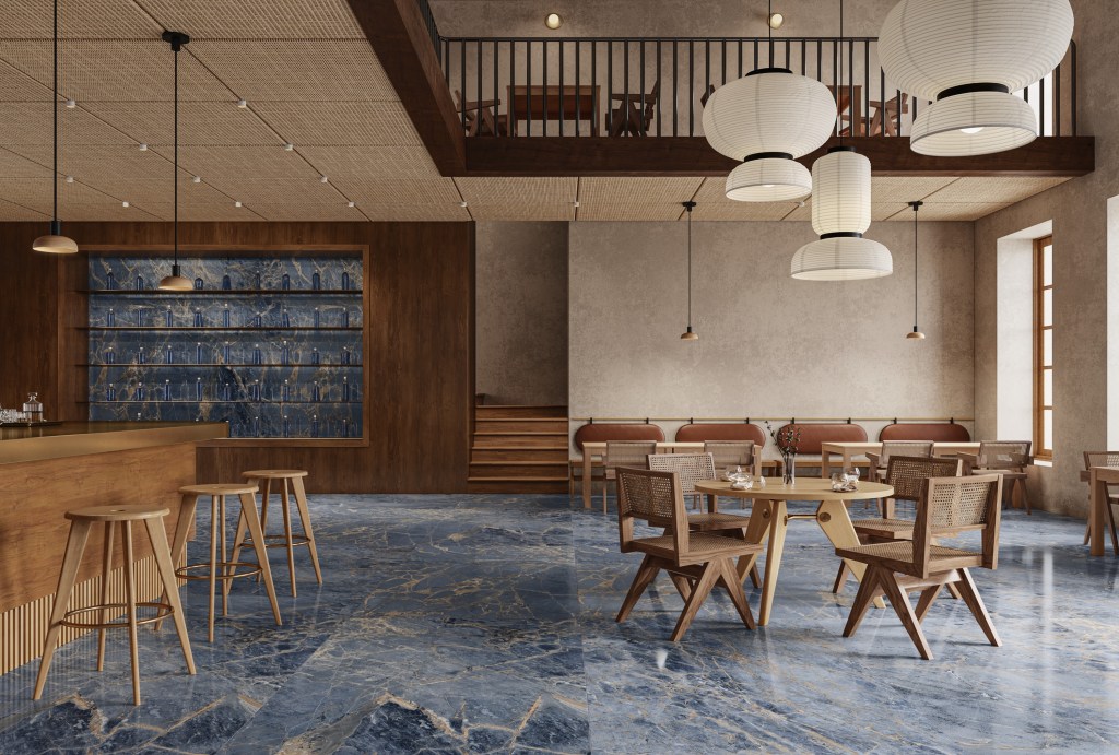 Foto mostra sala de jantar ampla com piso cerâmico em tom azulado que reproduz pedra, mesas e cadeiras de madeira.
