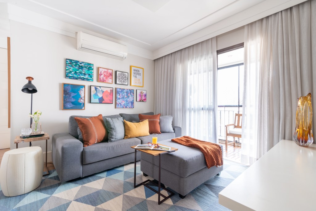 Sala de estar com varanda tem sofá cinza com almofadas coloridas, arranjo de quadros na parede, tapete grande geométrico e cortina.