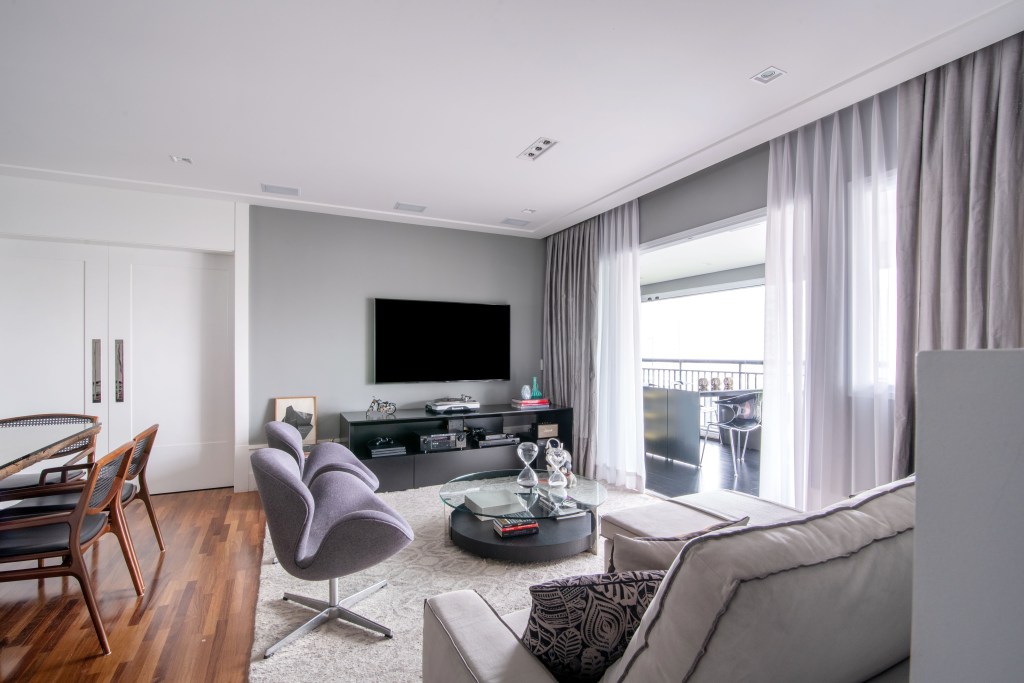 Sala de estar em apartamento com tapete, sofás, poltronas em tons de cinza claro, com rack e TV e pano de vidro aberto para varanda com cortina.