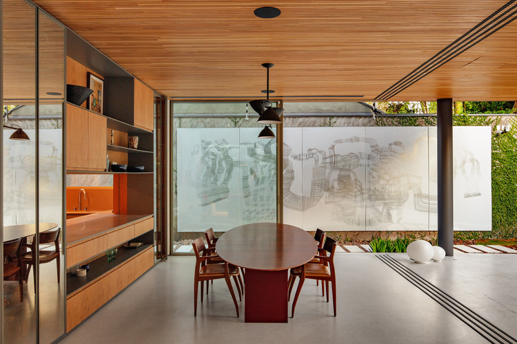 Sala de jantar com mesa de mesa oval e cadeiras de madeira, marcenaria e forro em madeira. Grandes painéis de vidro integram a sala ao jardim.