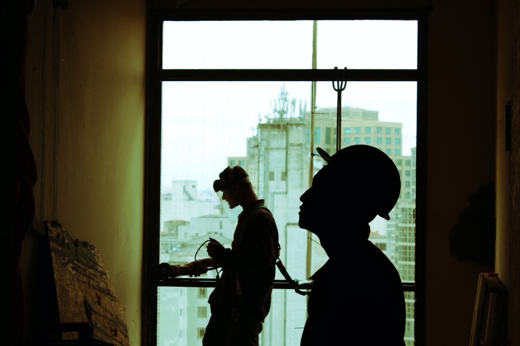 Dois trabalhadores da construção civil em contraluz próximos a uma janela. Um observa a parede e o outro usa uma máquina na esquadria da janela.