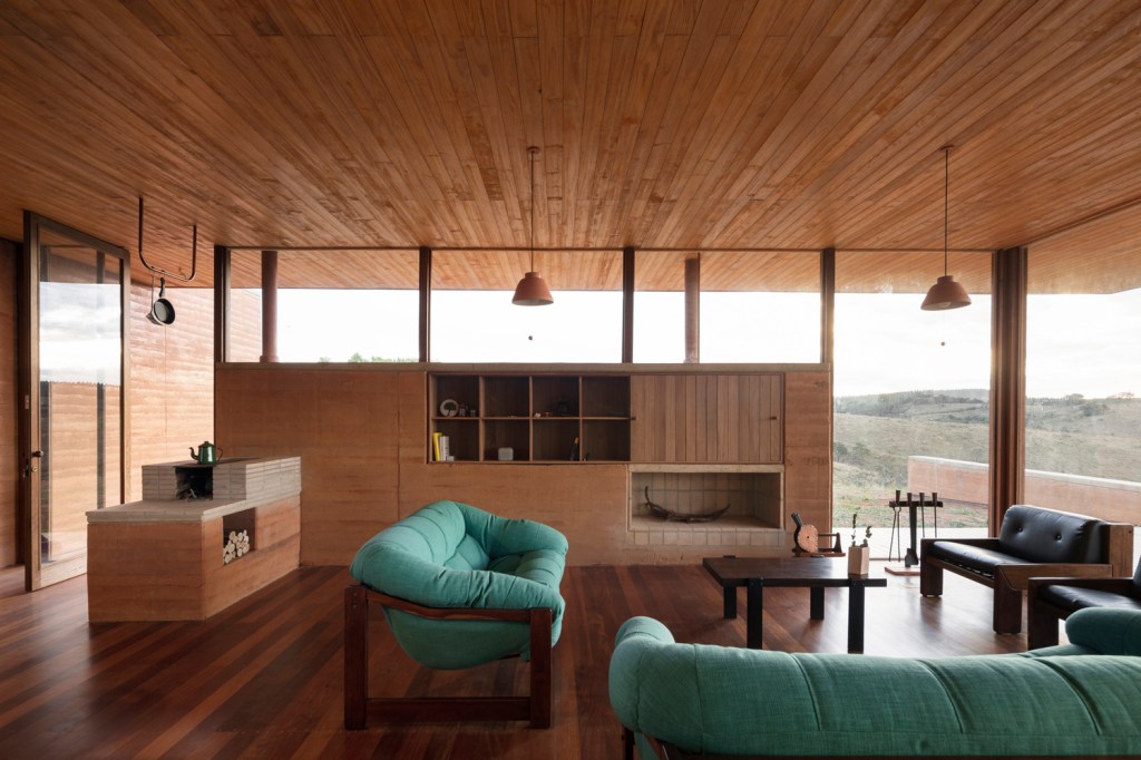 Sala de estar com lareira e fogão a lenha, piso e teto de madeira, paredes de terra e sofás em tecido azul.