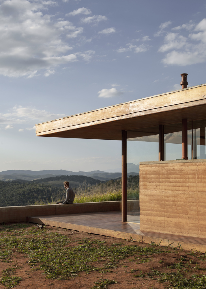 Detalhe de fachada de casa em taipa de pilão, com varanda e pessoa sentada à frente contemplando paisagem das montanhas.