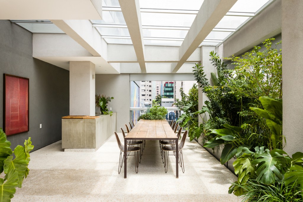 Mesa de jantar sob pergolado de concreto e vidro, em varanda de apartamento com um jardim tropical.
