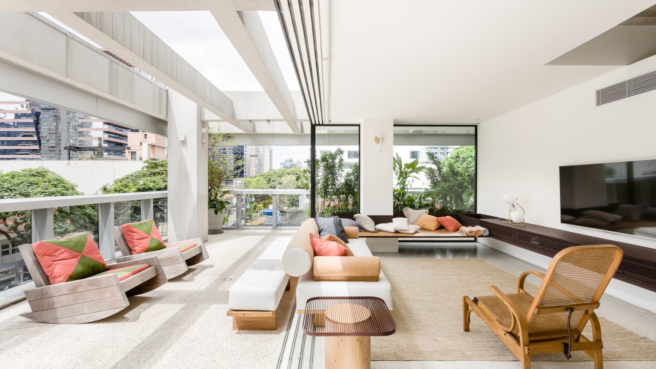 Sala de estar em apartamento tem varanda integrada e coberta com pérgula de vidro. Ambiente claro, com luz natural e móveis de madeira com almofadas coloridas.