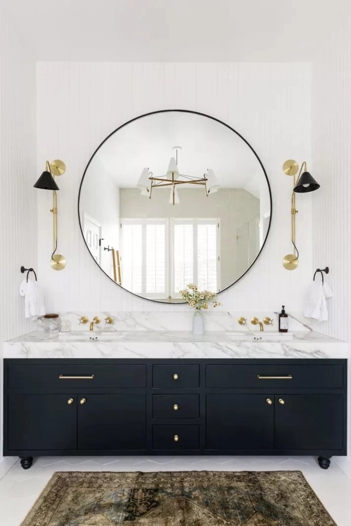 12 ideias fofas para decorar o banheiro-Cathie Hongs Banheiro com espelho circular, bancada de mármore, armários pretos e arandelas douradas com detalhes em preto.
