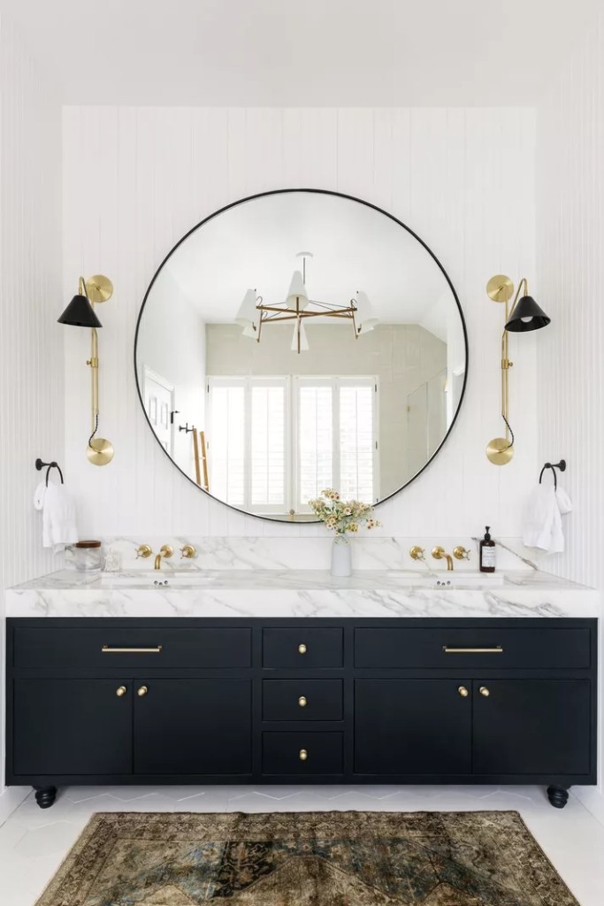 12 ideias fofas para decorar o banheiro-Cathie Hongs Banheiro com espelho circular, bancada de mármore, armários pretos e arandelas douradas com detalhes em preto.