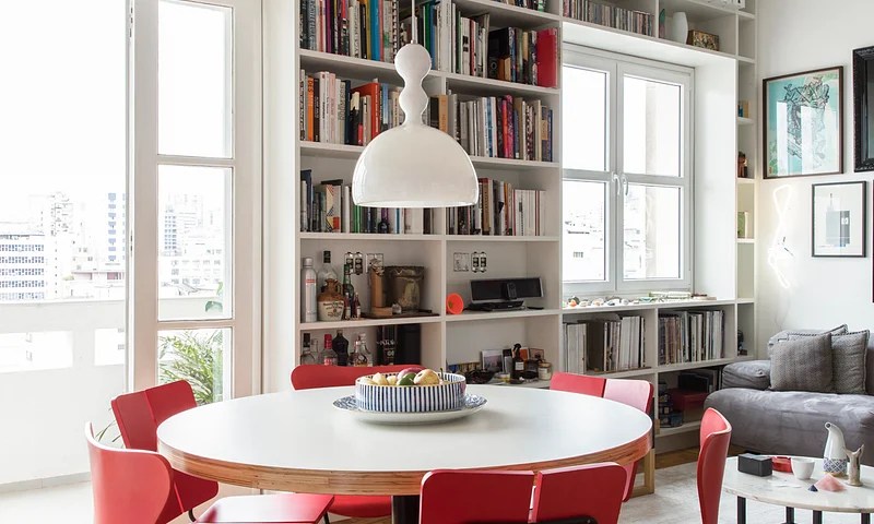 Sala de jantar com mesa redonda branca; estante com livros ao fundo; luminária