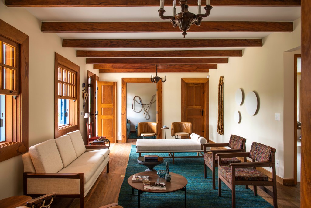 Sala de estar com vigas de madeira, sofá branco e cadeiras marrons e tapete turquesa
