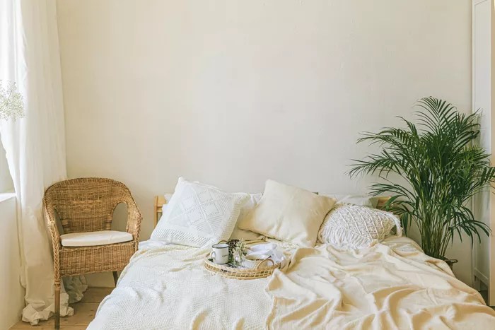 5-erros-comuns-de-quem-usa-o-estilo-Boho-Aleksandra Konoplia -EyeEm- Getty Images Cama com lençol e fronhas em tons de bege e branco. Em um lado, uma cadeira de vime, do outro uma planta.