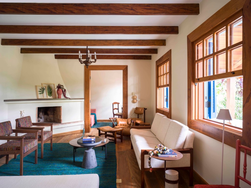 Sala de estar com vigas de madeira, sofá branco e cadeiras marrons e tapete turquesa