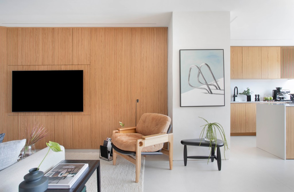 Sala de estar integrada à cozinha com painel de madeira, poltrona marrom de couro, obra de arte e mesinha de centro
