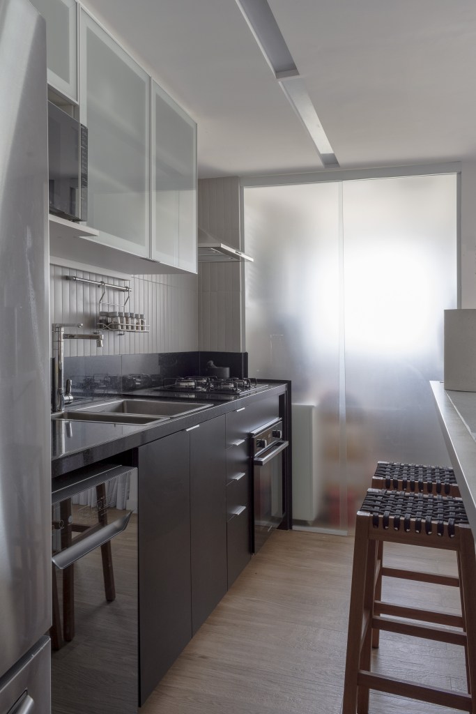 Cozinha estreita com móveis embutidos em estilo minimalista. A comunicação com a área de serviço é feita por uma porta de correr de vidro fosco.