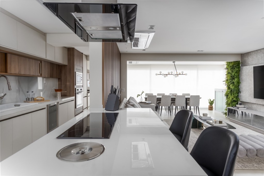 Foto mostra bancada de cozinha em revestimento branco, com cooktop, duas cadeiras de refeições e coifa. Ao fundo vemos a sala de jantar e uma varanda.