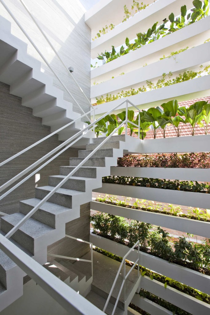 Foto mostra escada interna branca, com parede repleta de caixas de concreto que formam paredes verdes vazadas e cheias de vasos de plantas.