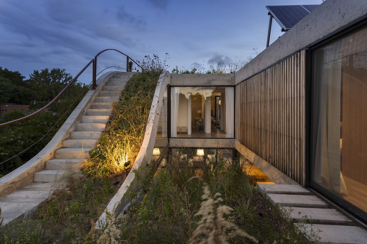 Foto mostra casa com escada que leva a uma cobertura ajardinada. Panos de vidro integram o interior ao jardim externo.