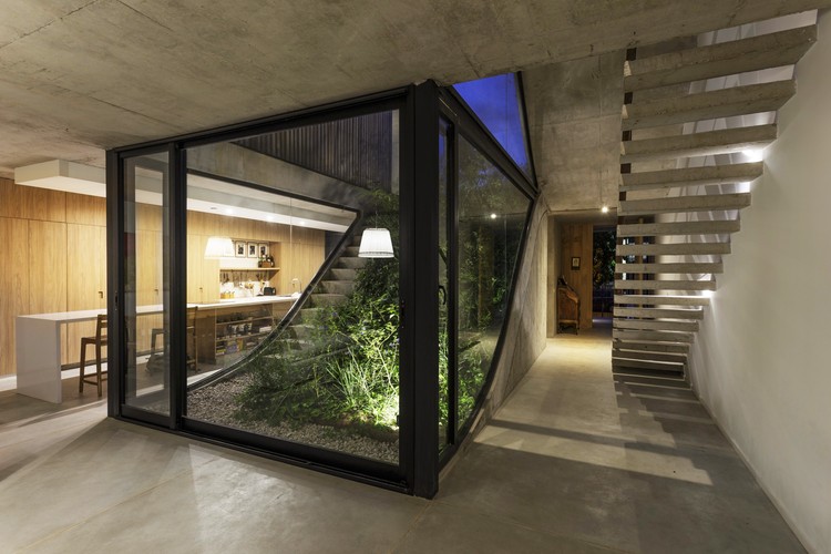 Caixa de estrutura metálica e fechamento em vidro conecta visualmente o jardim externo à área social da casa. Vê ainda uma escada de concreto.