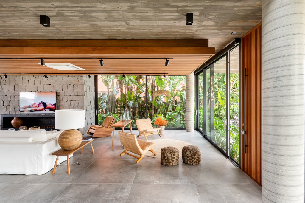 Foto mostra sala de estar com piso cimentício, totalmente integrada ao jardim externo por painéis de vidro em esquadrias metálicas.
