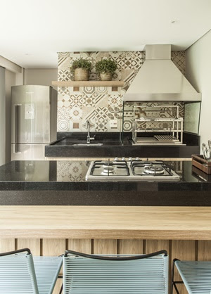 Foto mostra bancada de cozinha com cooktop e, atrás, bancada de trabalho com pia e churrasqueira com coifa, à frente de parede com ladrilhos coloridos em tons terrosos.
