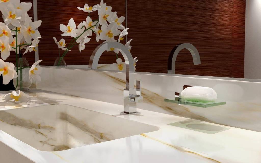 Foto mostra torneira de mesa em acabamento metálico, instalada sobre bancada de mármore em tom claro, com orquídea decorativa e saboneteira.