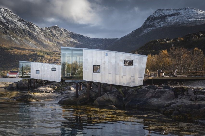 Foto mostra casa em formação rochosa à beira da água, com fachada geométrica em vidro e alumínio de tom acinzentado.