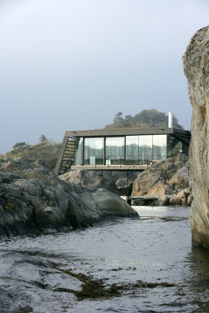 Foto mostra casa sobre encosta rochosa com fachada envidraçada e estrutura de concreto que se mimetiza com a natureza local.