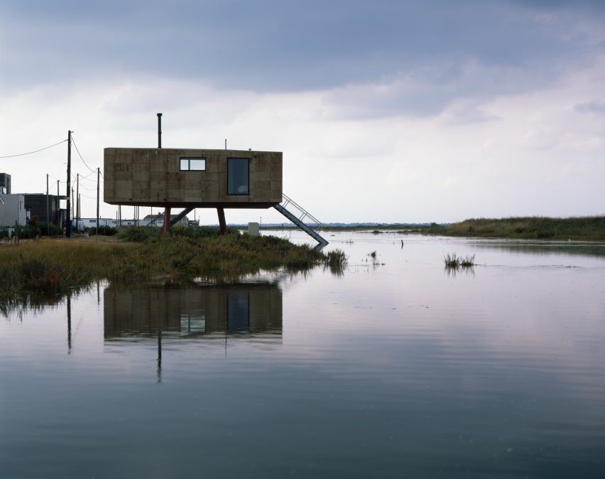 Foto mostra casa sobre palafitas à beira da água, que parece um lago.