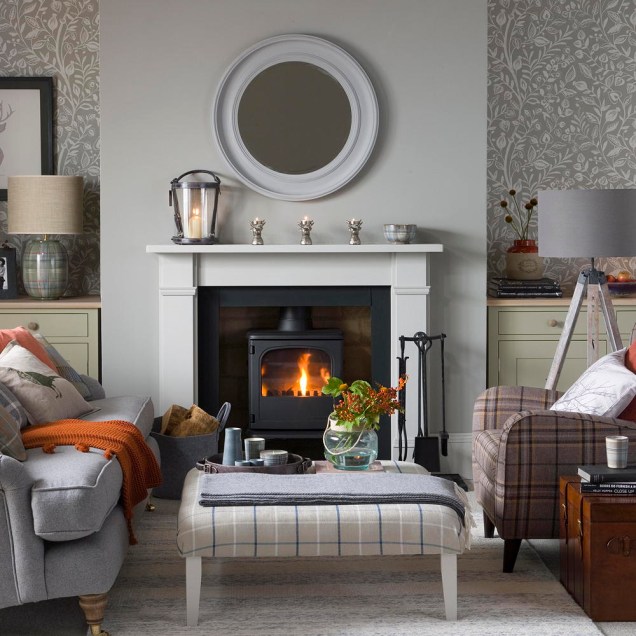 Grey modern country living room, nature pattern wallpaper, wood burning fireplace, lit fire, grey mantelpiece, grey sofa, plaid stool and armchair. Pub Orig