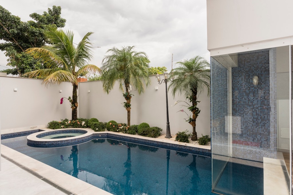 Imagem mostra área externa de uma casa com piscina de pastilhas azuis, borda de pedra natural e canteiro com buxinhos e palmeiras. Ao fundo, muro de proteção branco com arandelas para iluminar à noite.