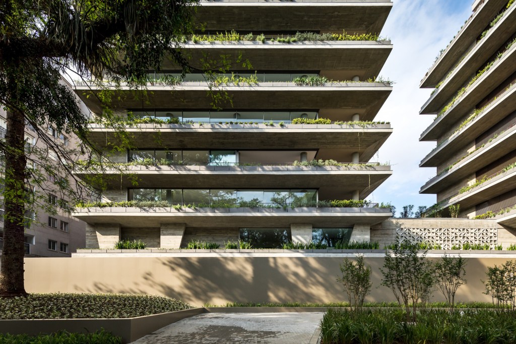Foto mostra fachada de condomínio vertical com duas torres que têm varandas cheias de plantas e jardins.
