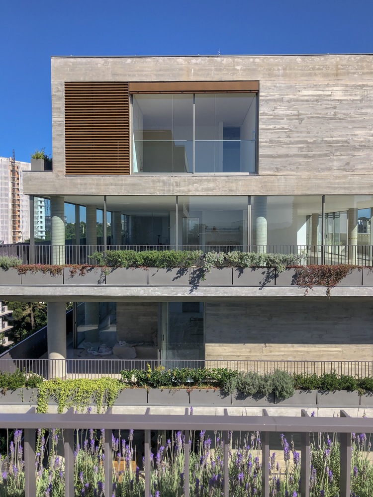 Foto mostra detalhe de fachada de apartamento com muitas plantas na varanda, janelas de vidro amplas e revestimento em concreto,