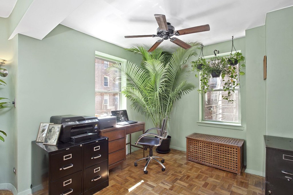 Home office de paredes verdes em que um canto de destaque é ancorado por uma grande planta que vai até o teto.