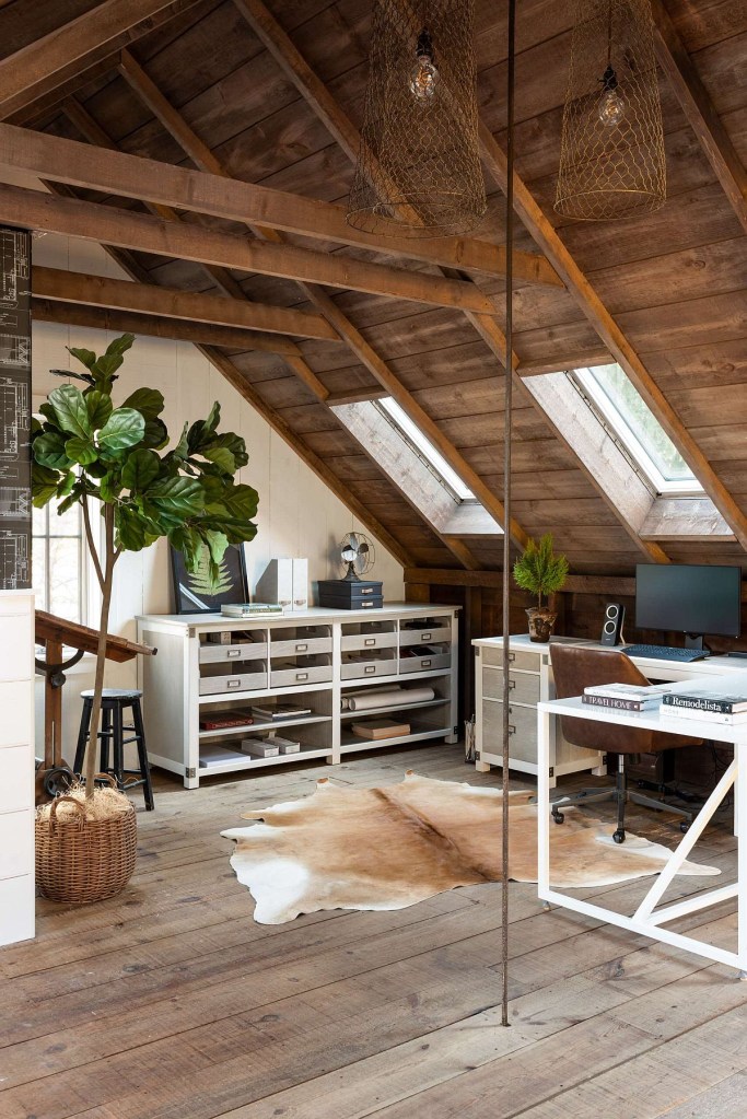 Sótão feito de home office. O teto é angulado, de madeira. Uma grande planta decora o ambiente.