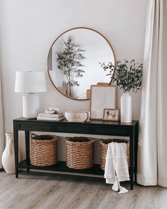 Hall de entrada com mesa de console preta decorada por cesta de vime embaixo e por cima um vaso de planta, abajur e quadros enquanto na parede de trás está um espelho redondo.