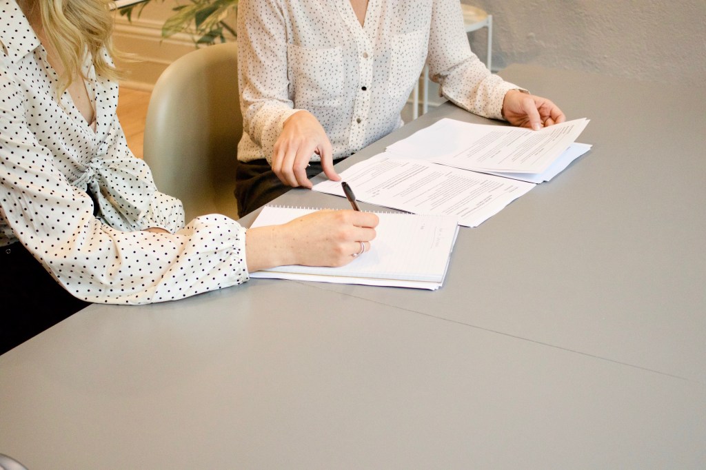 Foto mostra duas mulheres sentadas à mesa com papéis e uma delas escrevendo no papel, indicando uma assinatura de fechamento de negócio.