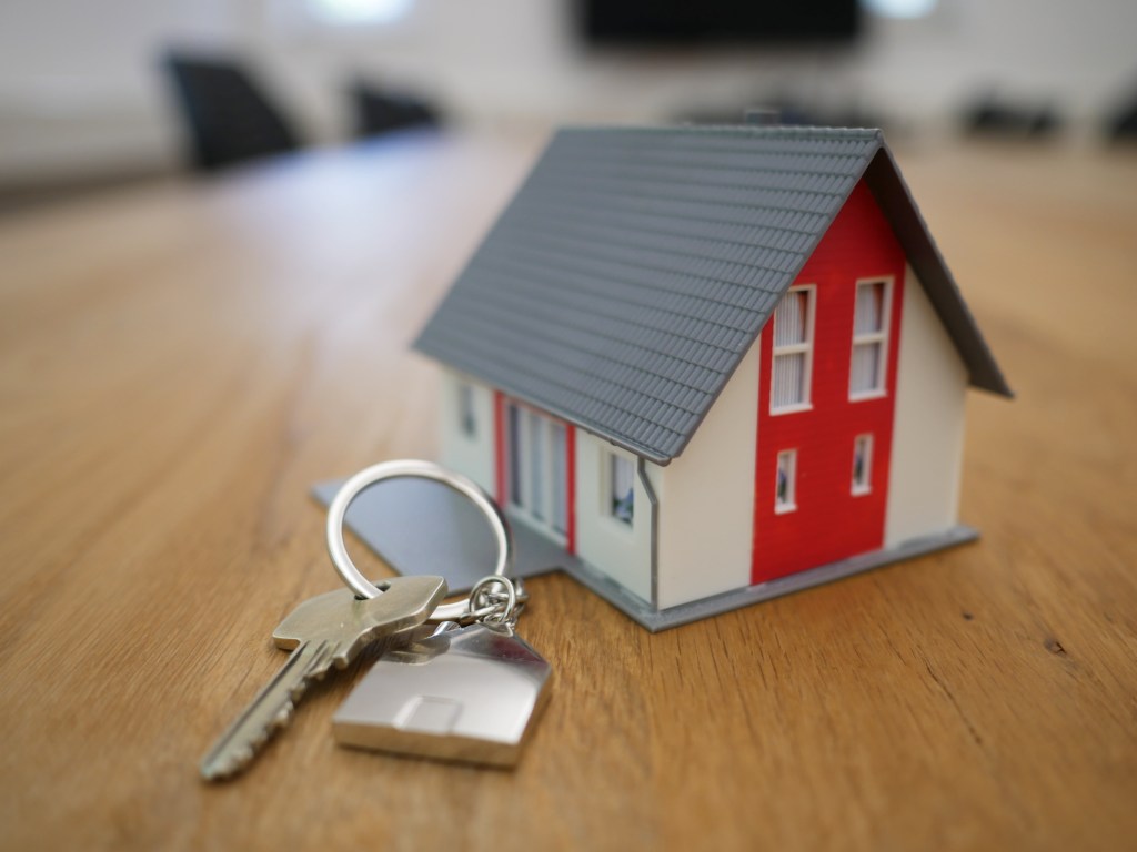 Foto mostra um chaveiro com uma casinha branca e vermelha e telhado cinza, com chave, sobre tampo de mesa de madeira.