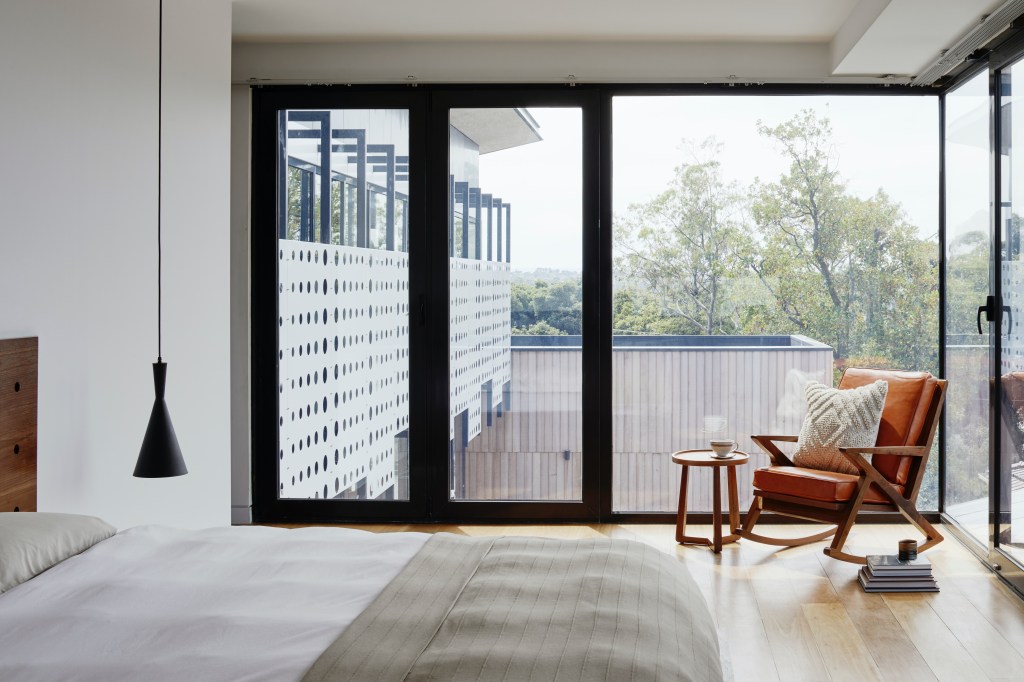 Foto mostra um quarto de casal com uma janela de vidro que toma toda a parede lateral da cama. Há uma poltrona de madeira e estofado laranja, com mesinha lateral perto da janela.