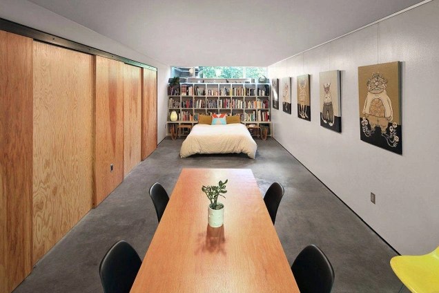 Foto mostra mesa de madeira com 4 cadeiras pretas em ambiente com piso de cimento queimado e, ao fundo, uma cama de casal e estante de livros revestindo toda a parede.